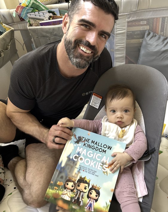 A dad smiling beside his baby in a bouncer, holding a hardcover picture book titled The Magical Cookies of the Mallow Kingdom