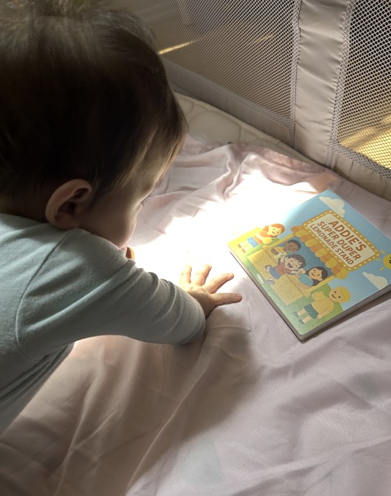 A baby on a play mat reaching toward a hardcover picture book titled Addie's Super Duper Lemonade Stand
