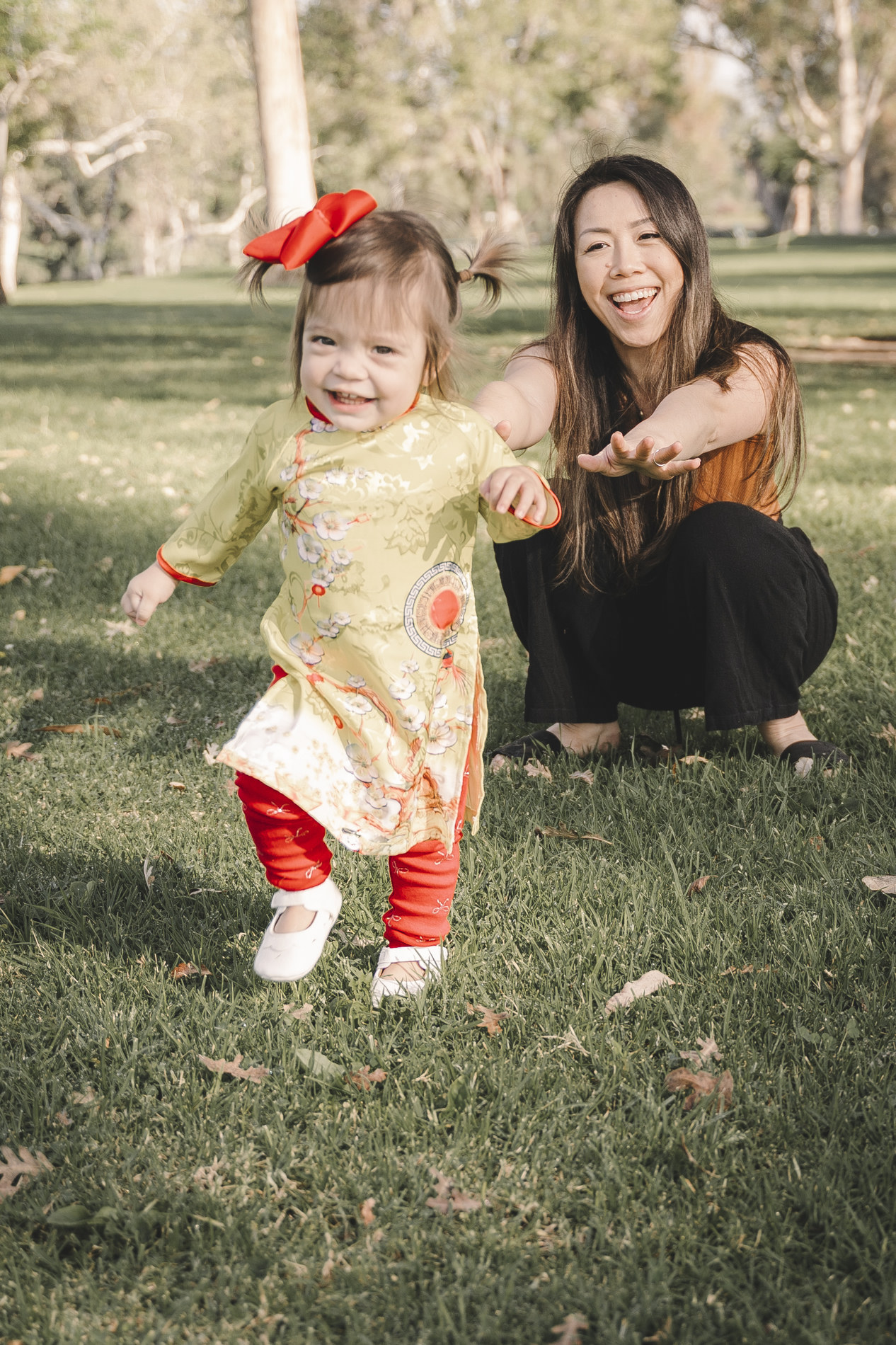 Julee kneeling on the grass, smiling, as her toddler in a yellow áo dài with a red bow walks toward the camera